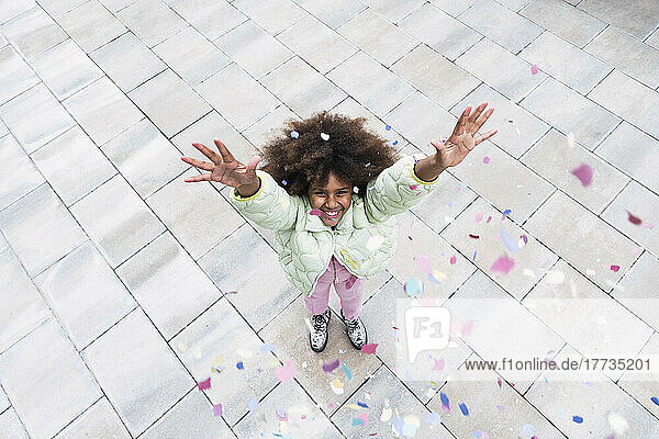 Fröhliches Afro-Mädchen mit erhobenen Armen steht inmitten von Konfetti  das auf den Fußweg fällt