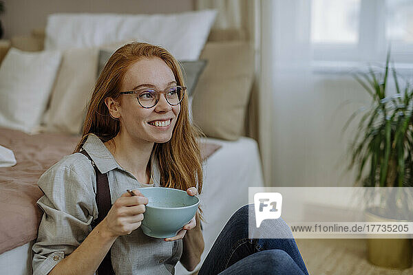 Happy woman with bowl of food sitting in front of bed at home