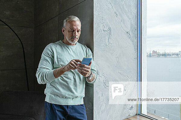 Mature man using mobile phone standing by window at home