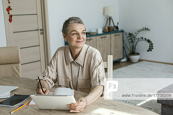 Nachdenkliche Frau sitzt zu Hause mit digitalem Tablet und Notebook am Tisch