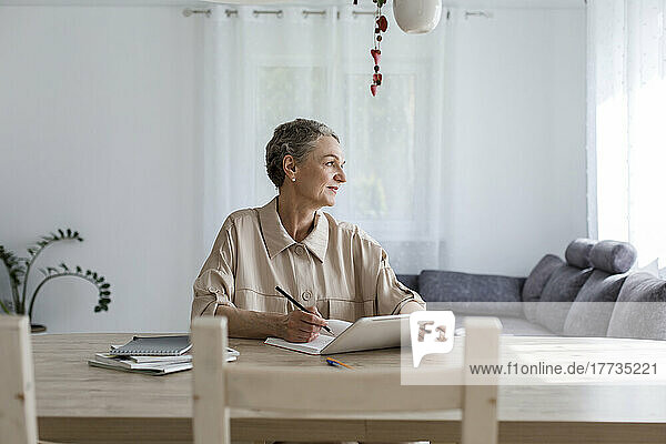 Thoughtful woman sitting at table at home with digital tablet and notebook