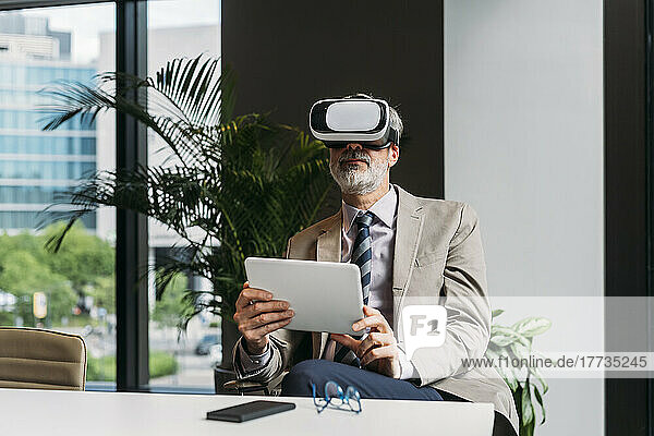 Businessman wearing virtual reality simulator sitting with tablet PC at desk in office
