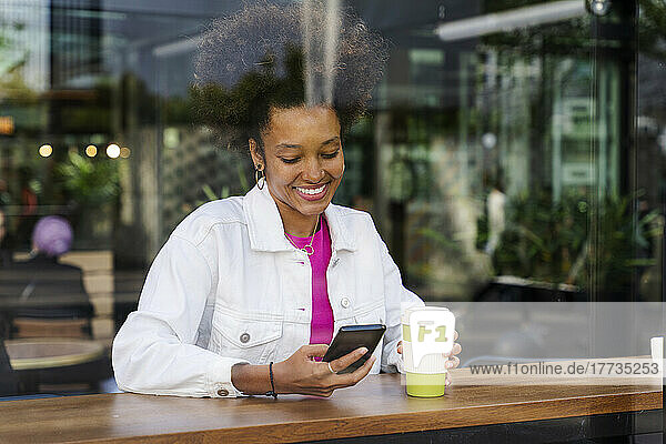 Smiling young woman with disposable coffee cup using mobile phone seen through glass