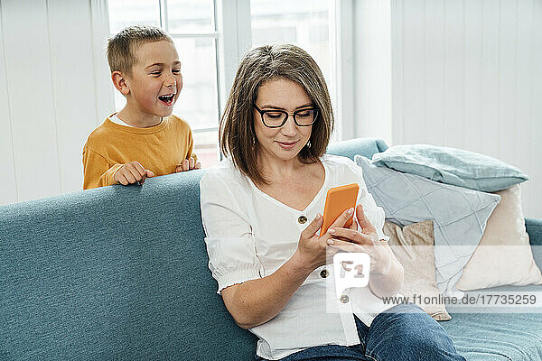 Happy boy looking at mother using smart phone on sofa at home