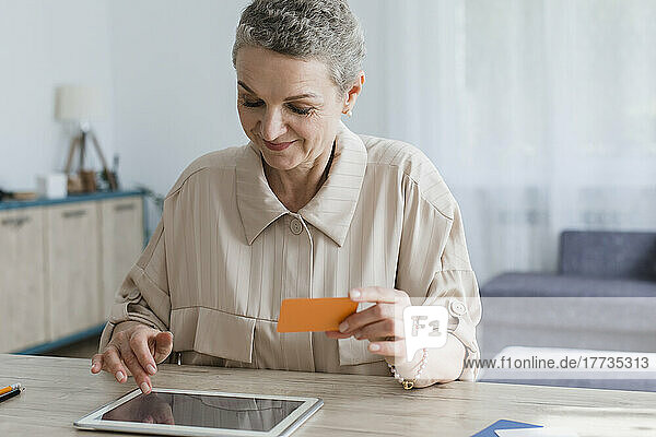 Woman sitting at table at home holding card and using digital tablet