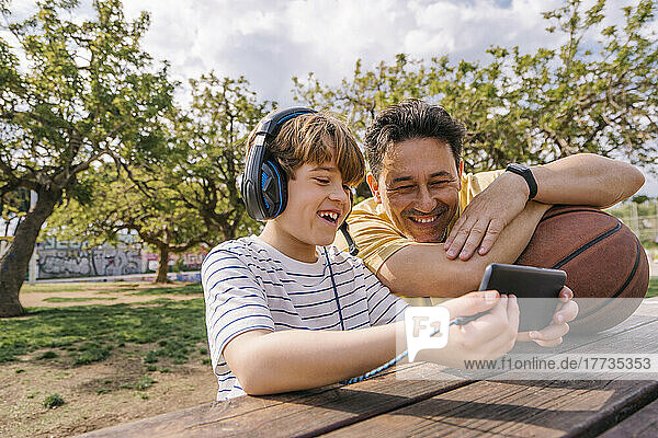 Smiling boy listening music through headphones sharing mobile phone with father sitting at table