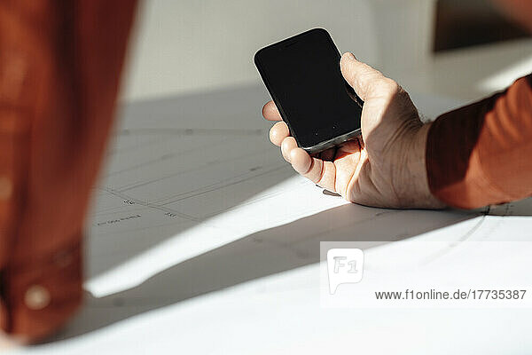 Hand of businessman holding smart phone on blueprint in office