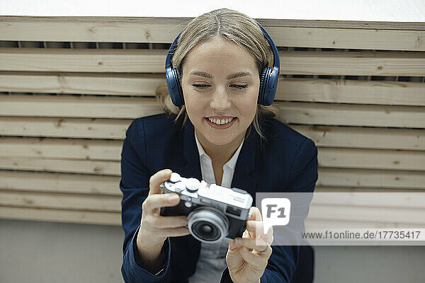 Lächelnde Geschäftsfrau hält Kamera in der Hand und hört im Büro Musik über drahtlose Kopfhörer