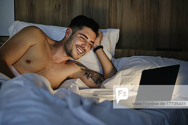 Happy man looking at laptop lying in bed at home