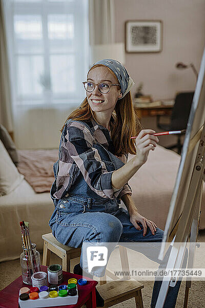 Happy woman with paintbrush sitting on stool by artist's canvas at home