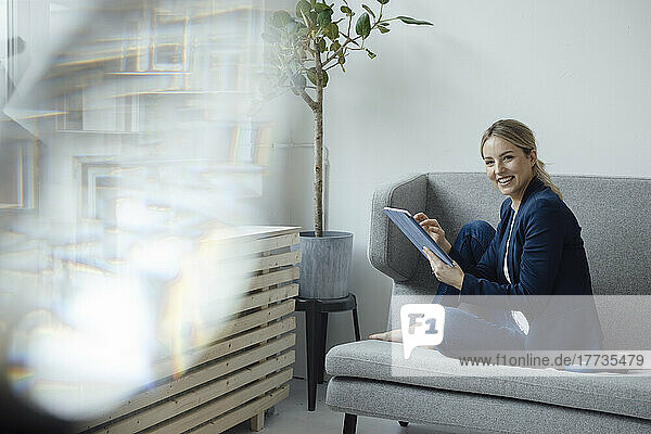 Smiling businesswoman holding tablet PC sitting on sofa in office