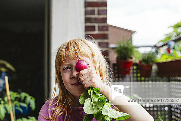 Girl covering eye with radish on balcony