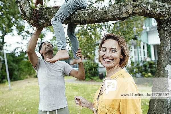 Happy woman standing in front of man looking at son sitting on tree at back yard