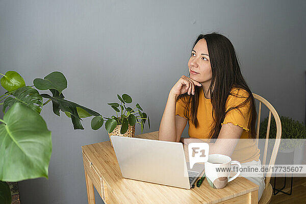 Thoughtful woman sitting with laptop and coffee mug at desk in home office