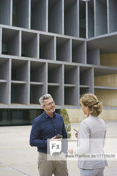 Smiling businessman holding mobile phone discussing with businesswoman in front of office building