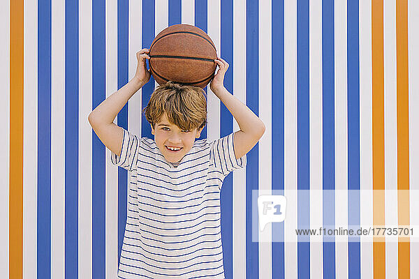 Happy boy holding basketball on head in front of orange and blue striped wall