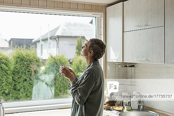 Mature woman holding tea glass at the window in kitchen
