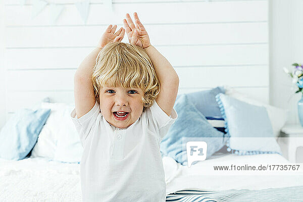 Cute boy with arms raised sitting on bed at home
