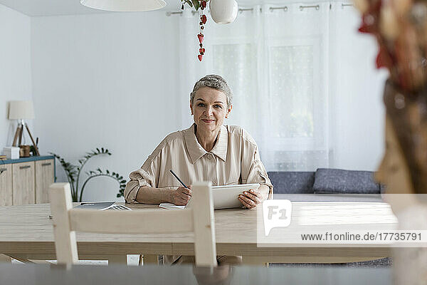 Portrait of woman using digital tablet and taking notes at table at home