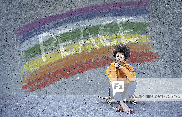Young woman sitting on skateboard in front of rainbow flag with text Peace