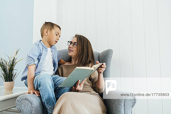 Boy reading book with mother sitting on armchair at home