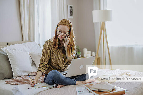 Smiling woman talking on mobile phone writing in book sitting with laptop on bed at home