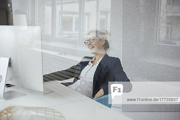 Happy businesswoman with desktop PC at desk seen through glass