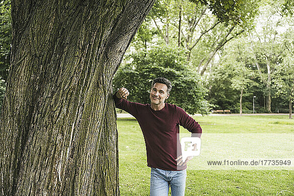 Happy mature man with hand on hip standing by tree at park