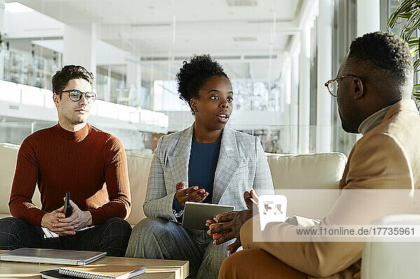 Junge Geschäftsfrau hält Tablet-PC in der Hand und diskutiert mit Geschäftsmann  der auf dem Sofa im Büro sitzt