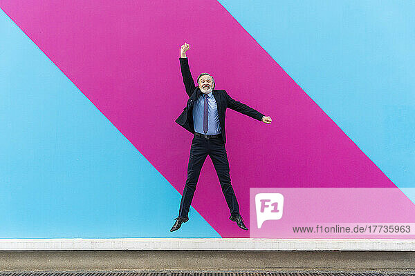 Glücklicher reifer Geschäftsmann springt mit erhobener Hand vor rosa und blauer Wand
