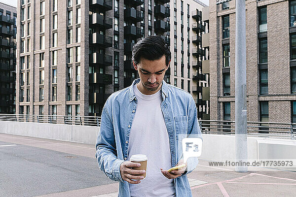 Man holding smart phone and disposable cup standing in front of building