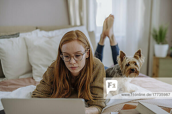Woman wearing eyeglasses using laptop lying by pet dog on bed at home
