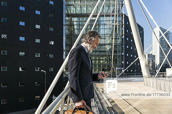 Happy mature businessman using smart phone standing by railing