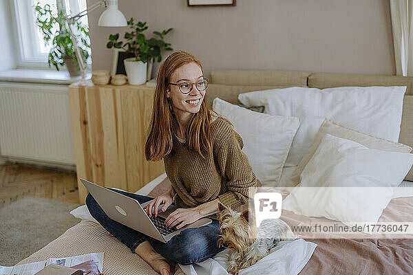 Glückliche Frau mit Brille sitzt mit Laptop neben Hund im heimischen Bett