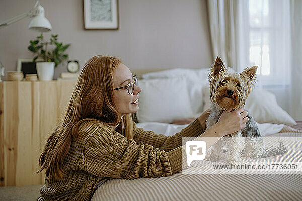 Smiling woman looking at pet dog sitting on bed at home