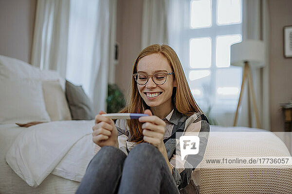 Happy redhead woman with pregnancy testing kit sitting in front of bed at home