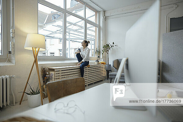 Businesswoman holding mobile phone sitting on window sill in office