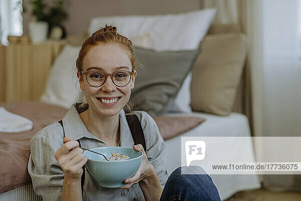 Smiling woman with bowl of food sitting in front of bed at home