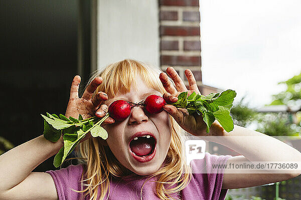 Girl with mouth open covering eyes with radish on balcony