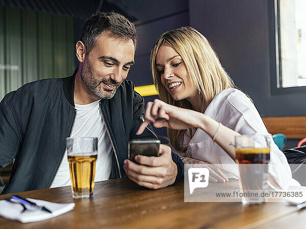Smiling woman pointing at smart phone held by man sitting at table in restaurant