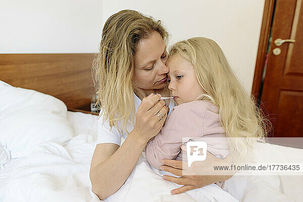 Mother carrying daughter and measuring temperature with thermometer in bedroom