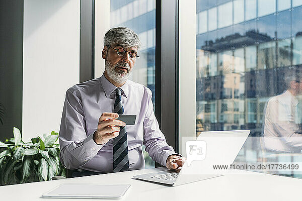 Businessman making payment with credit card through laptop sitting at desk in office