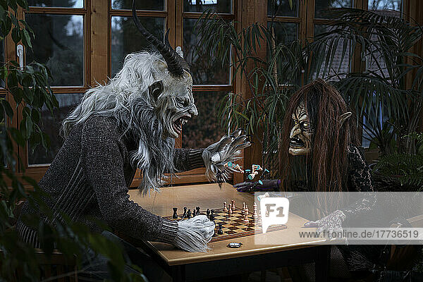 Angry man in spooky costume throwing chess piece on witch sitting at table