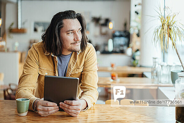 Smiling hipster man with tablet PC sitting at table in cafe
