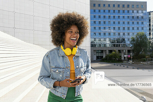 Cheerful Afro woman wearing wireless headphones standing with smart phone by steps on sunny day
