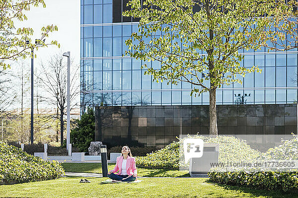 Geschäftsfrau sitzt an einem sonnigen Tag mit gekreuzten Beinen meditierend im Büropark