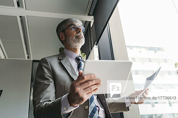 Mature businessman with documents and tablet PC standing by window in office