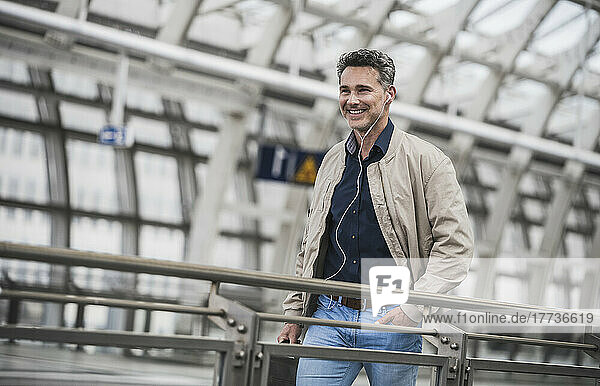 Happy mature man listening music through in-ear headphones walking with hand in pocket at railroad station