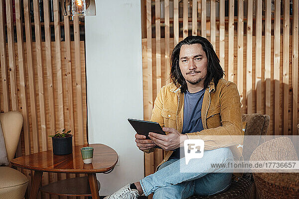 Lächelnder Hipster-Mann mit Tablet-PC sitzt auf einem Stuhl im Café