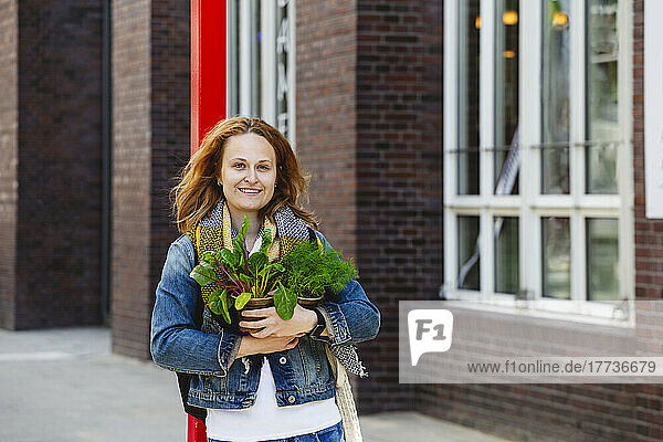 Smiling woman standing with dill and mangold plants outside building